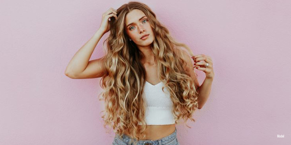 A confident woman with long, wavy hair posing against a pink wall, symbolizing evolving beauty standards.